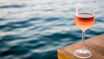 Ein Glas heller Rose auf einem Holzsteg über dem Meer. Unscharfe Wellen im Hintergrund.